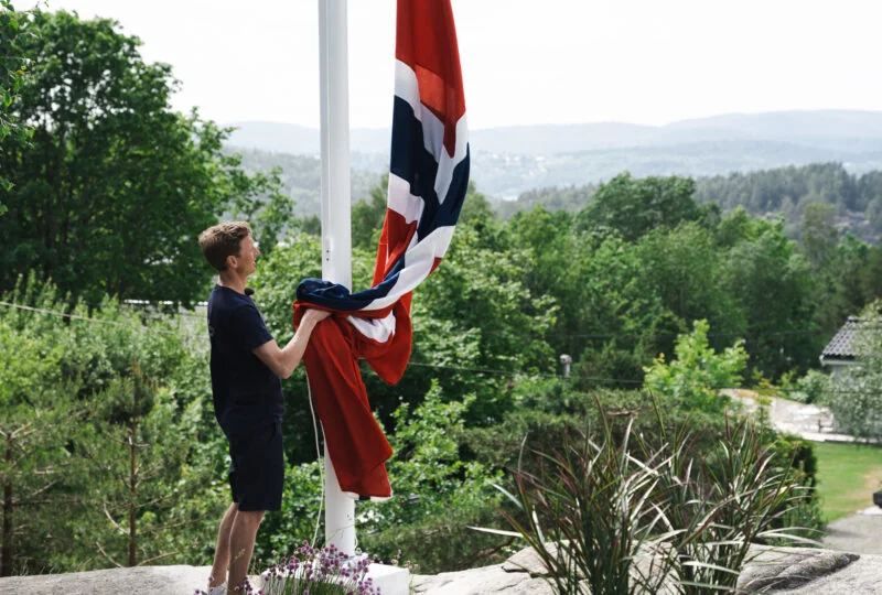 Ung mann heiser norsk flagg i hvit glassfiberflaggstang, med utsikt over skog og åser