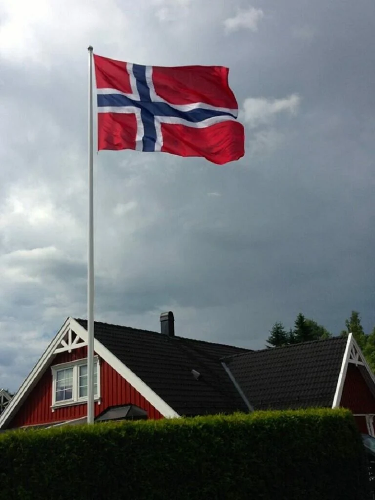 Norsk flagg i hvit flaggstang over et rødt hus med mørkt tak, mot skyet himmel
