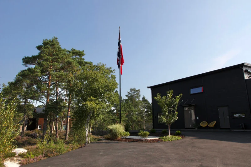Svart flaggstang i glassfiber med norsk flagg foran et svart hus blant furutrær